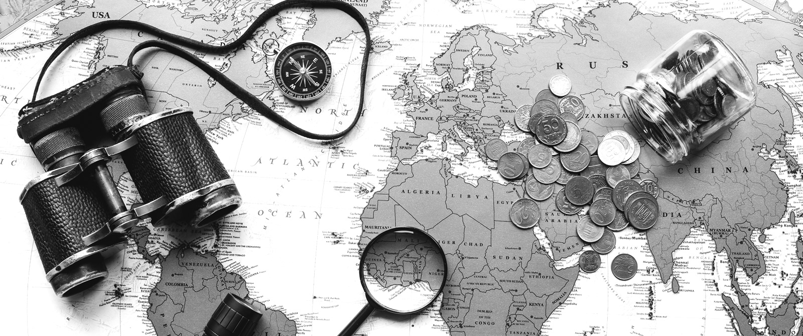 Black and white flat-lay of a world map with binoculars, a compass, a magnifying glass, and a jar of spilled coins, moody travel-planning atmosphere