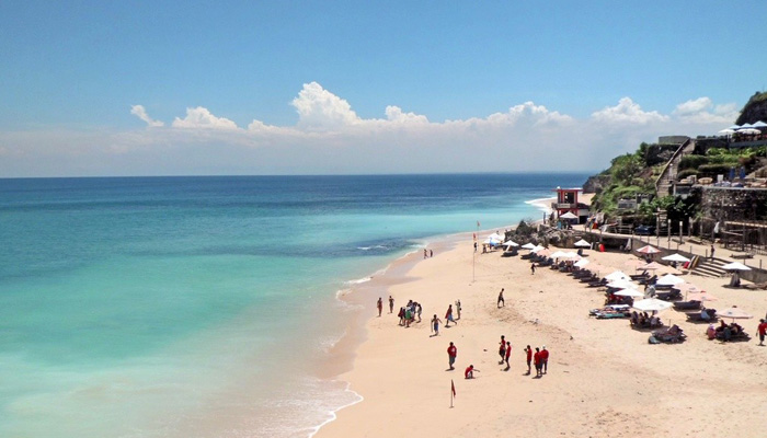 Aerial view of a sandy beach with turquoise water, scattered umbrellas and small figures, bright sunny mood