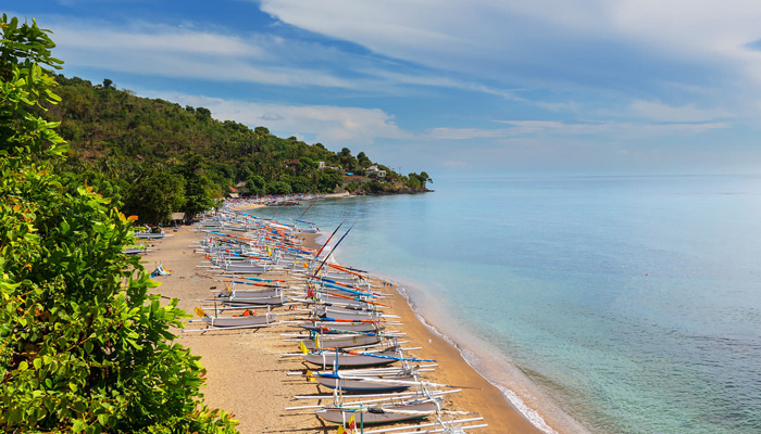 Rows of traditional outrigger boats lined on a sandy shoreline with green hills, calm clear-sky mood